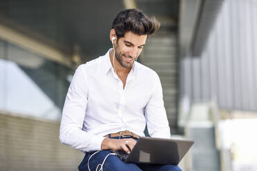 Smiling businessman wearing earphones using laptop outdoors in the city - JSMF01238
