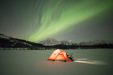 Fotograf in einem Zelt, beleuchtet von Nordlicht (Aurora Borealis) und Sternenhimmel in der Polarnacht, Svensby, Lyngen Alps, Troms, Norwegen, Skandinavien, Europa - RHPLF01415