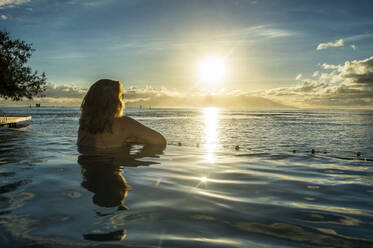 Frau genießt den Sonnenuntergang in einem Schwimmbad mit Moorea im Hintergrund, Papeete, Tahiti, Gesellschaftsinseln, Französisch-Polynesien, Pazifik - RHPLF01434