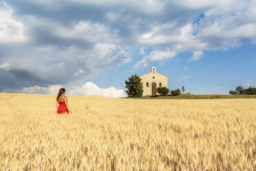 Frau im roten Kleid bewundert die Kapelle Notre-Dame-de-Sante in einem Weizenfeld, Entrevennes, Alpes-de-Haute-Provence, Provence-Alpes-Cote d'Azur, Frankreich, Europa - RHPLF01496