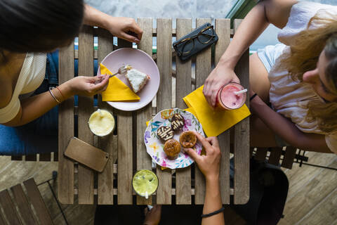 Draufsicht auf drei junge Frauen, die an einem Holztisch mit Smoothies und Keksen sitzen, lizenzfreies Stockfoto