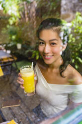 Porträt einer lächelnden jungen Frau, die ein gesundes Getränk hinter einer Glasscheibe hält - MGIF00682