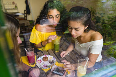 Drei junge Frauen mit Smoothies treffen sich in einem Café und teilen ihr Smartphone - MGIF00688