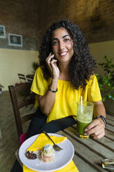 Porträt einer lächelnden jungen Frau, die mit einem Handy am Tisch sitzt und einen Smoothie trinkt - MGIF00695