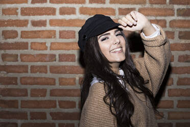 Portrait of smiling young woman wearing cap in front of brick wall - ABZF02449