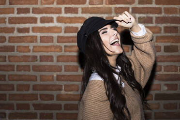 Portrait of laughing young woman wearing cap in front of brick wall - ABZF02450