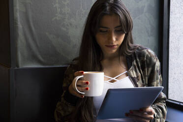 Junge Frau sitzt in einem Café mit einer Tasse Kaffee und benutzt ein Tablet - ABZF02461