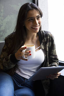 Junge Frau sitzt in einem Café mit einer Tasse Kaffee und hält ein Tablet in der Hand - ABZF02462