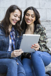 Happy young women watching tablet sitting on steps together in Madrid, Spain - ABZF02487