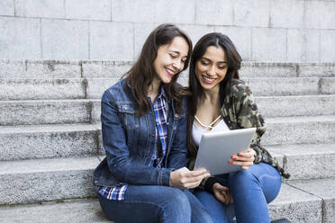 Happy young women watching tablet sitting on steps together in Madrid, Spain - ABZF02488