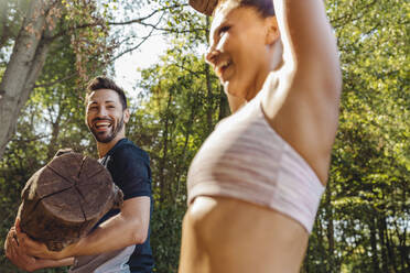 Mann beobachtet glückliche Frau, die schwere Baumstämme auf einem Fitnessparcours hochhebt - MFF04804