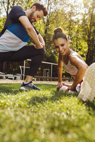 Mann und Frau dehnen sich im Gras in der Nähe eines Fitnessparcours, lizenzfreies Stockfoto