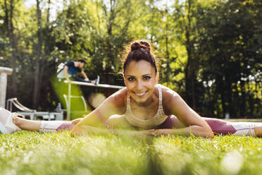 Porträt einer lächelnden Frau, die sich im Gras in der Nähe eines Fitnessparcours streckt - MFF04846