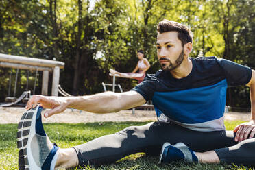 Mann dehnt sich im Gras in der Nähe eines Fitnessparcours - MFF04849