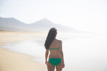 Back view of woman wearing green bikini standing on the beach looking at the sea, Fuerteventura, Spain - ABZF02524