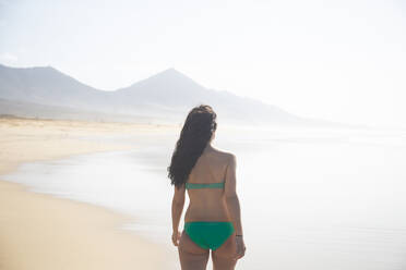 Rückenansicht einer Frau in einem grünen Bikini, die am Strand steht und auf das Meer schaut, Fuerteventura, Spanien - ABZF02524