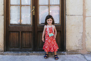 Porträt eines lächelnden kleinen Mädchens mit hölzerner Spielzeugkamera im roten Kleid mit Blumenmuster - GEMF03111