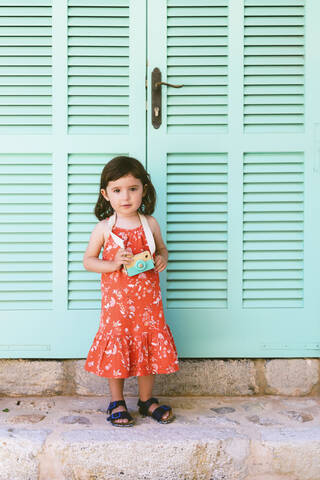 Porträt eines kleinen Mädchens mit hölzerner Spielzeugkamera im roten Kleid mit Blumenmuster, lizenzfreies Stockfoto