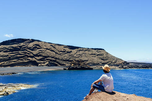 Mann sitzt auf einer Klippe und blickt auf den Strand El Golfo, Lanzarote, Kanarische Inseln, Spanien - KIJF02626