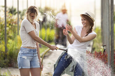 Zwei glückliche junge Frauen haben Spaß beim Blumengießen mit dem Schlauch im Gewächshaus - JSRF00556