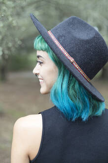 Lächelnde junge Frau mit gefärbten blauen und grünen Haaren und Hut an einem regnerischen Tag - JPTF00278
