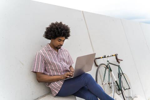 Young man sitting on steps and using laptop, bicycle leaning on a wall stock photo