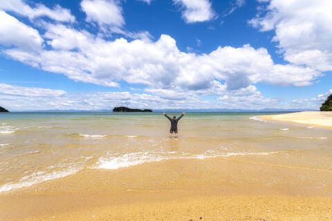 Rückansicht eines reifen Mannes, der mit erhobenen Armen im Meer steht, vor einem bewölkten Himmel am Abel Tasman Coastal Track, Südinsel, Neuseeland, lizenzfreies Stockfoto