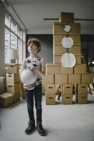Porträt eines rothaarigen Jungen mit Globus, der vor einer aus Pappkartons gebauten Rakete steht, lizenzfreies Stockfoto