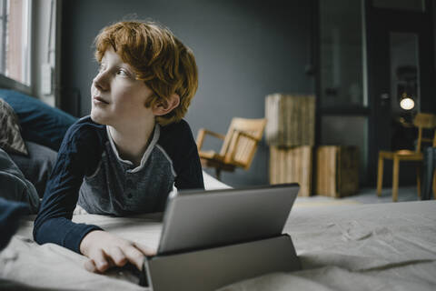 Redheaded boy lying on couch with digital tablet looking out of window stock photo