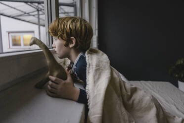 Sad boy leaning on window sill looking out of window in the evening - KNSF06243