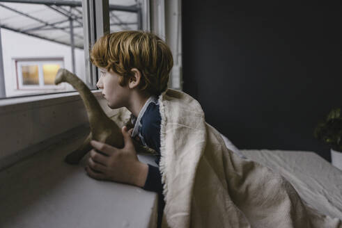 Sad boy leaning on window sill looking out of window in the evening - KNSF06243