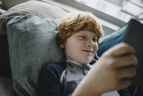 Porträt eines lächelnden rothaarigen Jungen, der auf einer Couch liegt und ein digitales Tablet benutzt - KNSF06249