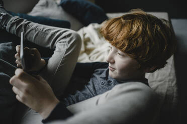 Redheaded boy lying on couch using digital tablet - KNSF06250