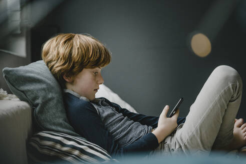 Rothaariger Junge liegt auf der Couch und schaut auf sein Handy - KNSF06254