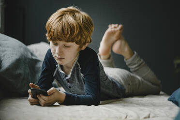 Portrait of redheaded boy lying on couch looking at mobile phone - KNSF06255