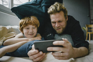 Portrait of father and son lying on couch looking at cell phone - KNSF06262