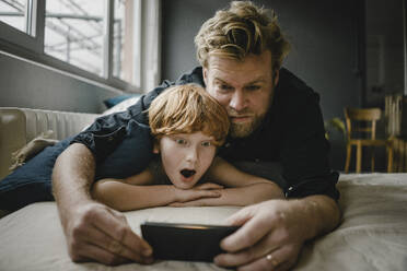 Portrait of father and son lying on couch looking at cell phone - KNSF06263