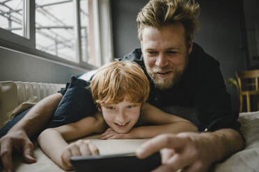 Portrait of father and son lying on couch looking at cell phone - KNSF06264