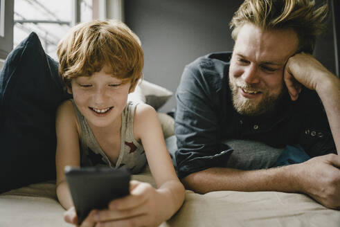 Porträt eines glücklichen Vaters und eines Sohnes, die zusammen auf der Couch liegen und auf ihr Handy schauen - KNSF06266