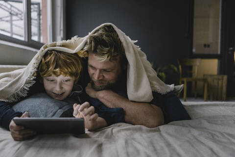 Vater und Sohn liegen zusammen unter einer Decke und schauen auf ein digitales Tablet, lizenzfreies Stockfoto