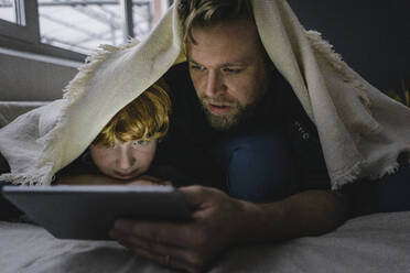 Father and son lying together under blanket looking at digital tablet - KNSF06292