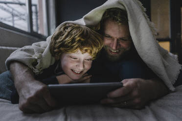 Father and son lying together under blanket looking at digital tablet - KNSF06293
