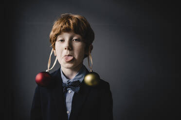 Porträt eines rothaarigen Jungen mit Fliege und Weihnachtskugeln, die die Zunge herausstrecken - KNSF06304