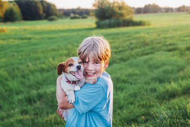 Portrait of happy boy carrying dog on field during sunny day, Poland - MJF02432