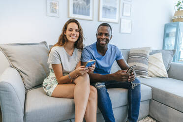 Portrait of smiling couple in living room with cell phones - KIJF02662