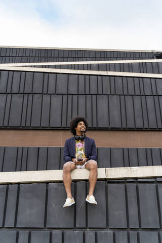 Stilvoller Mann mit Handy an einer Wand sitzend, lizenzfreies Stockfoto