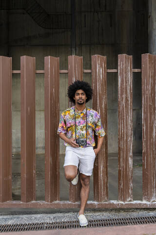 Man with a camera wearing colorful shirt leaning against a fence stock photo