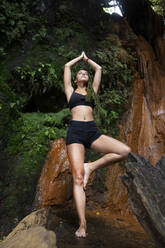 Young woman practising yoga at waterfall, tree position - LJF00714