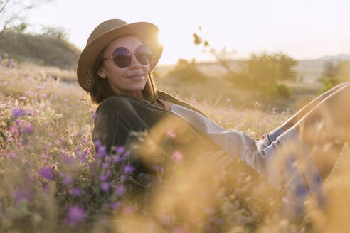 Junge Frau mit Sonnenbrille und Sonnenhut, die bei Sonnenuntergang auf einer Wiese sitzt - KNTF03080