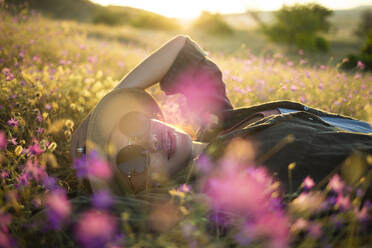 Junge Frau mit Sonnenhut auf einer Wiese liegend bei Sonnenuntergang - KNTF03082